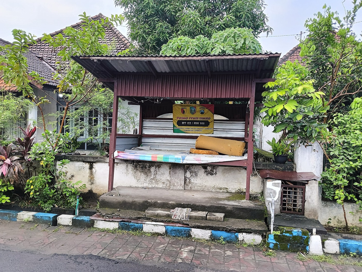 Pos ronda di tepi gang dengan papan nama RT 07 RW 04 di bawah atap seng